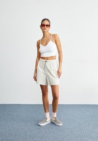 Biały prążkowany crop top na cienkich ramiączkach, beżowe szorty ze sznurkiem i czarno-białe buty slip-on w kratkę. Okulary przeciwsłoneczne z czerwonymi szkłami.