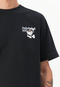 Svart t-skjorte med kort erme i bomull med et hvitt grafisk trykk av "Daily Grind NYC" og en tegneseriefigur på venstre bryst.