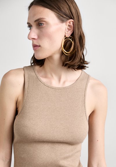 Donna con capelli corti castani, che indossa grandi orecchini a cerchio dorati e un top senza maniche beige a coste, che guarda verso sinistra.