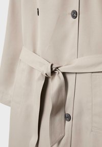 Beige trenchcoat med et bundet bælte i taljen, der har sorte knapper, to sidelommer og et glat, tekstureret stof.