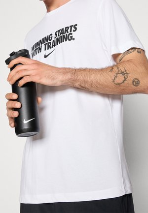 Personne portant un t-shirt blanc Nike avec le texte "Winning starts with training", tenant une bouteille d'eau noire Nike, montrant des tatouages sur son bras.