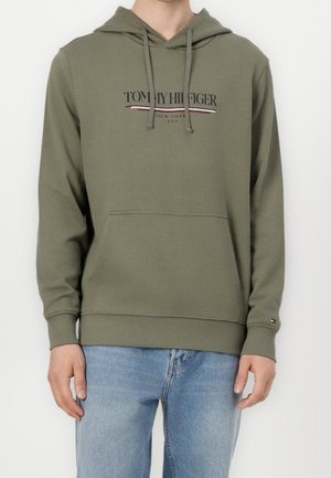 Mann trägt olivgrünen Tommy Hilfiger Hoodie mit Fronttasche und hellblauer Jeans, steht vor einfachem weißen Hintergrund.