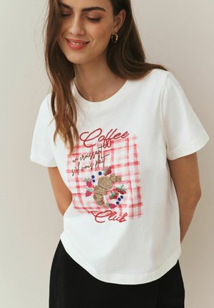 Weißes Baumwoll-T-Shirt mit einem rosa karierten Stickdesign, das ein Croissant und florale Akzente zeigt. Rundhalsausschnitt und kurze Ärmel.