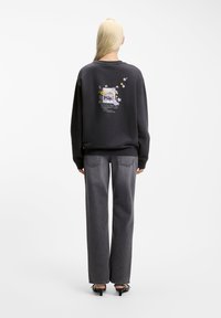 Άτομο με μακριά ξανθά μαλλιά φοράει μαύρο φούτερ με floral σχέδιο και κείμενο στην πλάτη, σε συνδυασμό με γκρι τζιν και μαύρες γόβες, στραμμένος προς τα πίσω.