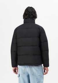 Sort pufferjakke med høj krave og vandret syninger. Materialet ser syntetisk og glat ud, kombineret med let blå slidte jeans.