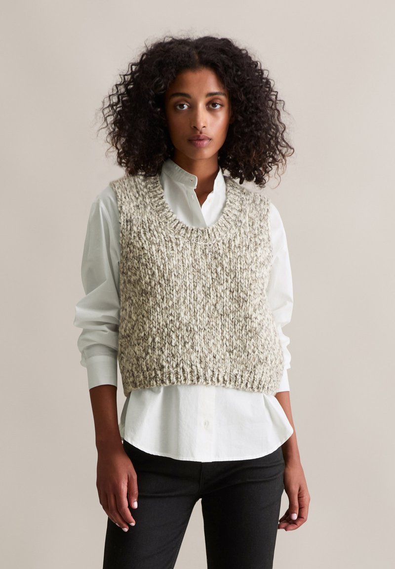 Veste tricotée avec un col large et un ourlet côtelé, réalisée en fil beige et gris. Portée sur une chemise blanche boutonnée avec un pantalon noir.