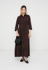 Femme debout portant une longue robe boutonnée à motifs noir et rouge foncé, tenant un sac à main noir et portant des chaussures plates rouges foncées.