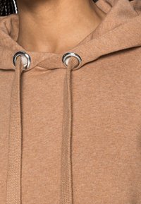 Detalle de sudadera con capucha marrón con ojales plateados y cordones gruesos, con una tela texturizada y un escote relajado.