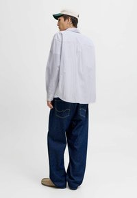 Person iført en hvid stribet skjorte, brede mørkeblå jeans, beige sko og en hvid og grøn baseballcap, der står med ryggen til.