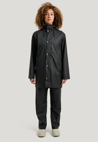 Musta, vedenpitävä sadetakki, jossa on nepparit ja kiristettävä huppu, yhdistettynä yhteensopiviin housuihin ja vaaleansävyisiin kengiin.