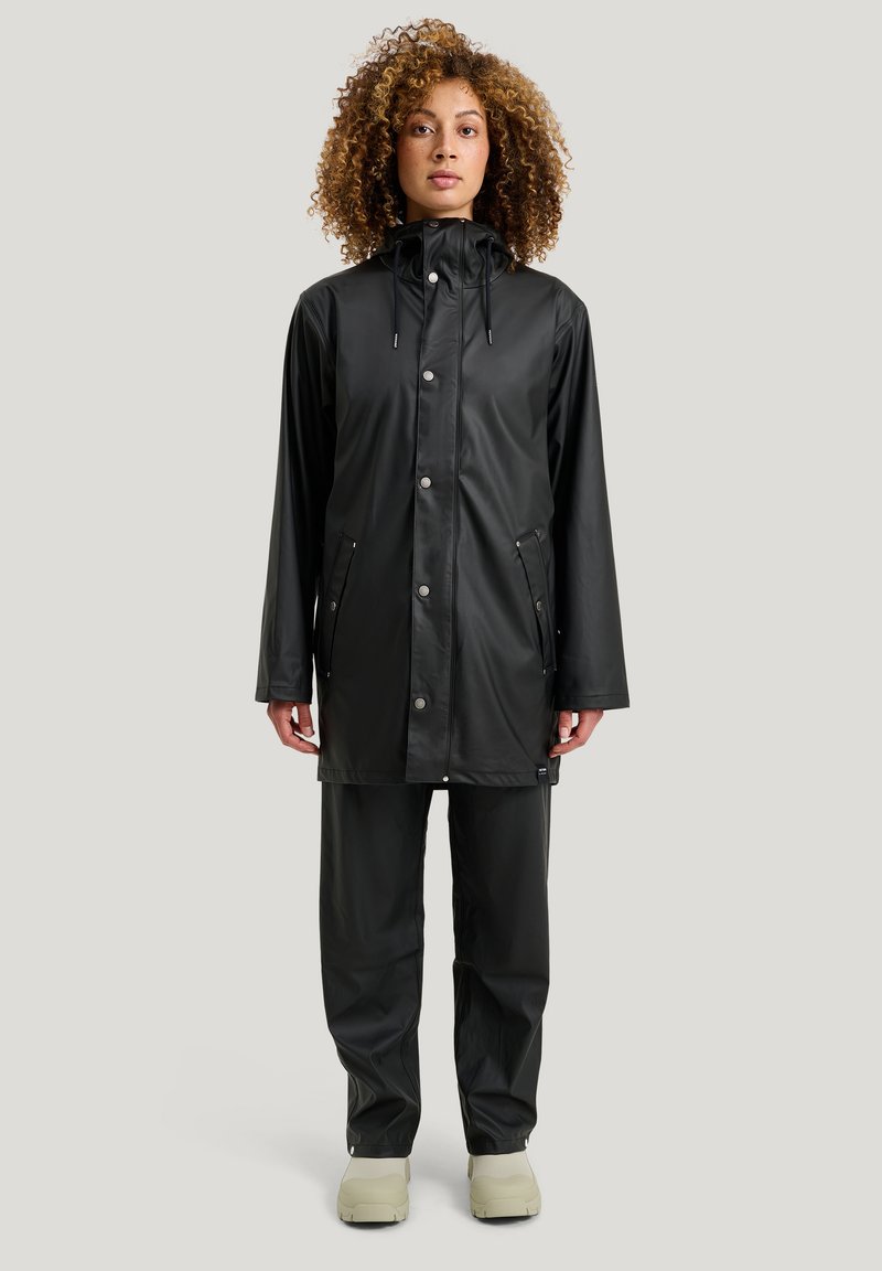 Musta, vedenpitävä sadetakki, jossa on nepparit ja kiristettävä huppu, yhdistettynä yhteensopiviin housuihin ja vaaleansävyisiin kengiin.