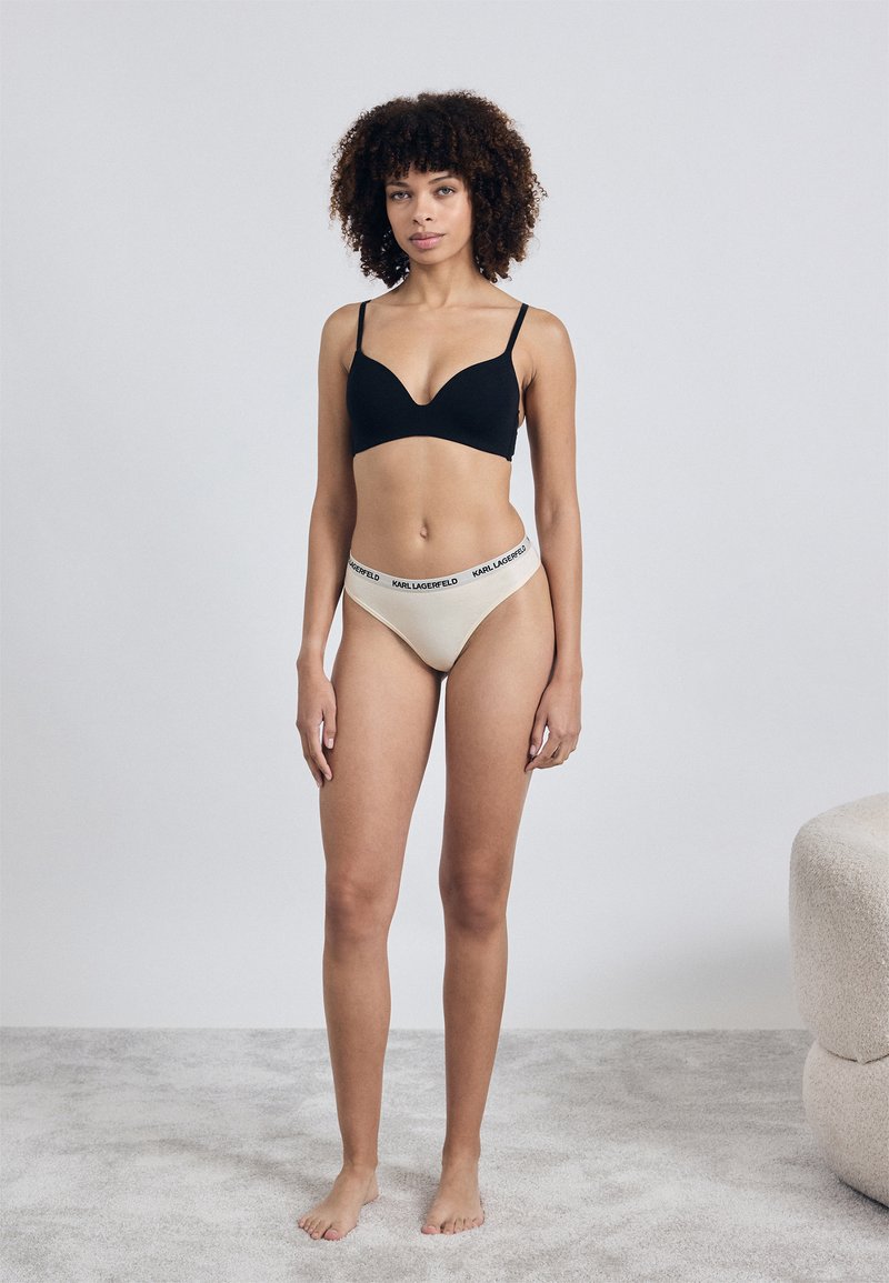 Reggiseno nero imbottito con spalline regolabili, abbinato a mutande color crema con un elastico in vita con logo nero. Texture liscia, design semplice.