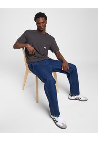 Dark gray cotton t-shirt with left chest pocket, blue denim jeans, and white sneakers with gray accents, sitting on a wooden chair.
