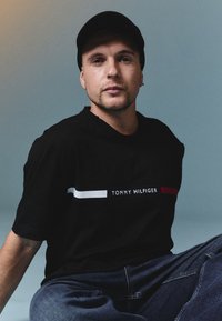 Crna pamučna majica s okruglim izrezom, s vodoravnom bijelo-crvenom prugom i natpisom "TOMMY HILFIGER" izvezenim bijelom bojom sprijeda.