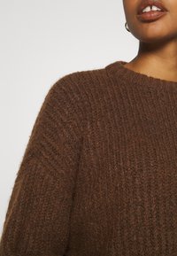 Zbliżenie na osobę ubraną w grubą, brązową sweter, pokazującą część szyi, ramienia i warg z złotymi kolczykami w kształcie koła.