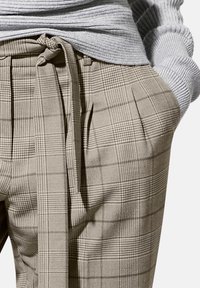 Beige geruite broek met een ceintuur in de taille, voorzien van plooien en een gestructureerde stof. Het patroon bevat kleine en grote vierkanten.