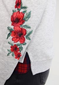 Sudadera gris con bordado floral rojo en las mangas, con un acabado texturizado y aberturas laterales. Debajo, se puede ver una camisa de cuadros negra y roja.