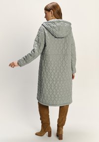 Hellgrüner, gesteppter Mantel mit Kapuze, der eine lange, lockere Passform, ein strukturiertes Diamantmuster und Taschen an den Seiten aufweist. Getragen mit beige Kniehochstiefeln.