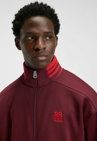 Giacca zip-up bordeaux con colletto alto, dettagli a strisce rosse e logo ricamato sul petto. Tessuto liscio e design sportivo.