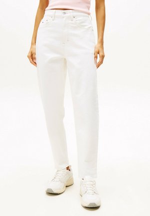 Persoon draagt witte jeans met een hoge taille, lichtroze top en witte sneakers, staand tegen een effen witte achtergrond.