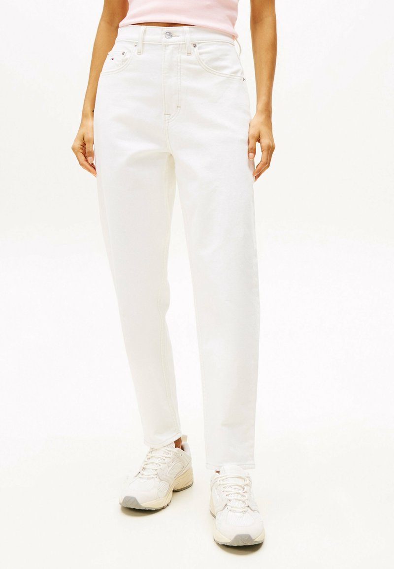 Personne portant un jean blanc taille haute, un top rose clair et des baskets blanches, debout devant un fond blanc uni.
