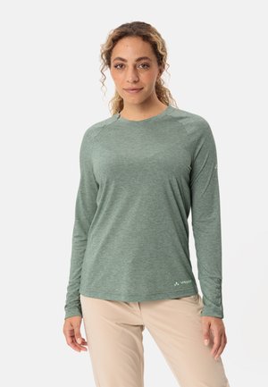 Langsleeve groene prestatie t-shirt met een textuurafwerking, raglanmouwen en een logo op de onderzijde, gecombineerd met lichtgekleurde broek.