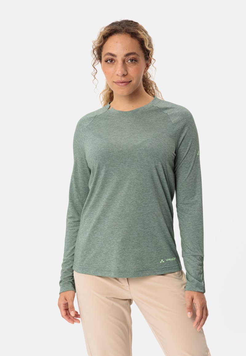 Langärmliges grünes Performance-Shirt mit strukturierter Oberfläche, Raglanärmeln und einem Logo auf der unteren Vorderseite, kombiniert mit hellen Hosen.