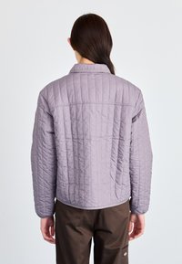 Μπουφάν από καπιτονέ ύφασμα σε λιλά απόχρωση με γιακά, μακριά μανίκια και ευθεία γραμμή. Διαθέτει απαλή υφή και ορατές ραφές.