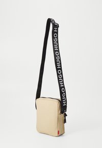 Borsa a spalla quadrata beige in tessuto, con tracolla nera decorata da logo bianco, chiusura con zip e tasca laterale.