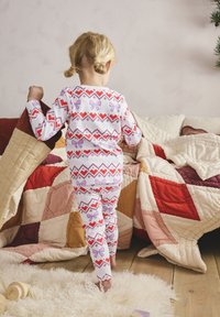 White pajamas featuring red hearts and pink bows in a geometric pattern, paired with a child carrying a decorative stocking in a cozy setting.