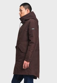Homme de profil portant un long manteau à capuche brun foncé avec des poches avant et le logo Aigle sur la manche.