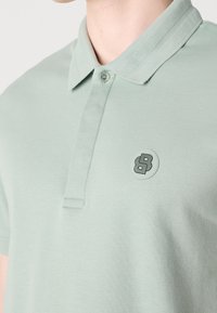 Polo de color verde menta hecho de suave algodón, con cuello acanalado, tapeta con botones y un parche de logo negro en el pecho.