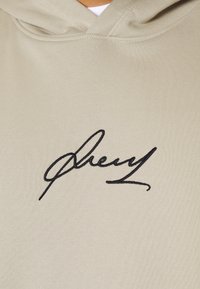 Felpa beige con una firma nera ricamata sul davanti. Il tessuto è liscio, con una tasca a marsupio standard e un cappuccio regolabile.