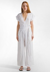 Kvinna med långt rött hår som bär en vit linnesparkdräkt med omlindning och svarta sandaler, stående mot en enkel vit bakgrund.