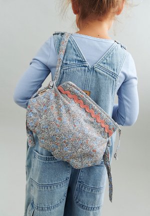 Kind draagt lichtblauw shirt met lange mouwen en spijkeroverall, en heeft een schoudertas met bloemenpatroon en roze golvende rand op de rug.