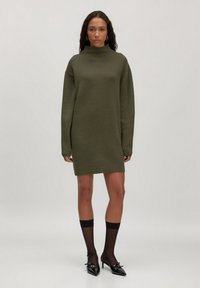 Olivengrünes, übergroßes Strickkleid mit hohem Kragen, langen Ärmeln und gerader Silhouette, kombiniert mit schwarzen Strumpfhosen und spitzen High Heels.