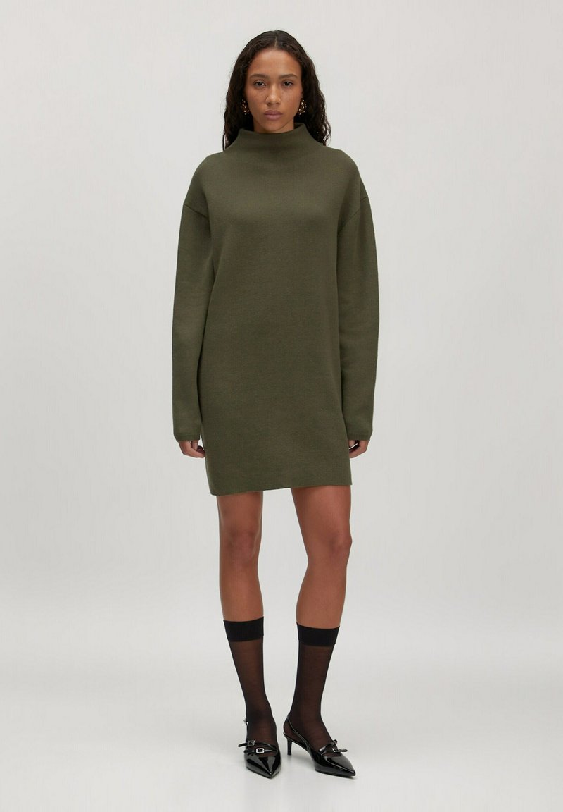 Olivengrünes, übergroßes Strickkleid mit hohem Kragen, langen Ärmeln und gerader Silhouette, kombiniert mit schwarzen Strumpfhosen und spitzen High Heels.