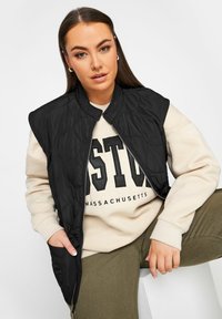 Svart vadderad ärmlös väst med ståkrage, dragkedjestängning och sidofickor, lager-på-lager över en krämfärgad sweatshirt med stora svarta bokstäver.