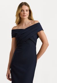 Vállról lelógó, tengerészkék ruha hajtogatott mellrésszel, szűk szabással és sima szövet textúrával, oldalsó varrással.