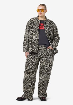 Oversize-Jacke und -Hose im Zebra-Design aus beige-schwarzem Stoff, getragen über einem grauen T-Shirt mit rotem Text. Gelbe Sonnenbrille vervollständigt das Outfit.