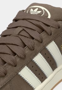 Baskets en daim marron avec trois bandes en cuir blanc, lacets texturés et un logo sur la languette. Présente des accents de surpiqûres en zigzag.