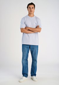 T-shirt azzurro a maniche corte con taschino frontale, abbinato a jeans di denim di lavaggio medio e sneakers bianche, in piedi su uno sfondo neutro.