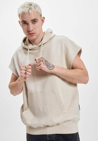 Beige ärmelloses Kapuzensweatshirt mit einer Fronttasche, einem Kordelzug und dem Text "NINETEEN NINETY" in Weiß. Weiche Stofftextur.