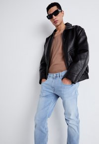 Chaqueta de cuero negra con acabado texturizado, parte superior beige ajustada y jeans de mezclilla azul claro. Unas gafas de sol cuadradas negras añaden un acento moderno.