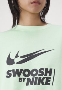 Sweat-shirt vert clair avec un grand logo Swoosh Nike noir et le texte "SWOOSH BY NIKE" en lettres majuscules et en gras. Tissu doux au toucher.