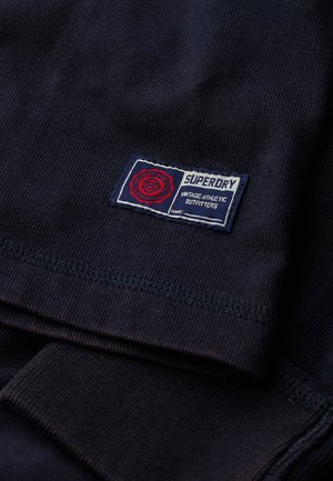 Navy blue cotton t-shirt with ribbed texture and a Superdry logo patch featuring red, white, and blue colors on the bottom hem.