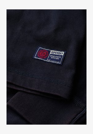 Granatowa bawełniana koszulka z prążkowanym wzorem i naszywką z logo Superdry w czerwonym, białym i niebieskim kolorze na dole.