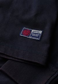 Marineblaues T-Shirt mit gerippter Textur, versehen mit einem Superdry-Label in Rot und Weiß. Das Label beinhaltet die Wörter "vintage athletic outfitters".