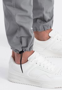 Šedé užší joggingové kalhoty s elastickými manžetami, spárované s bílými teniskami s perforovanými detaily a plochými gumovými podrážkami.