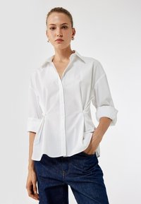 Valkoinen nappipaita rentoa istuvuutta, jossa on kaulus ja käännetyt hihat. Sujuva kangas, sivuilla rypytyksiä, yhdistettynä tummiin denim-housuihin.